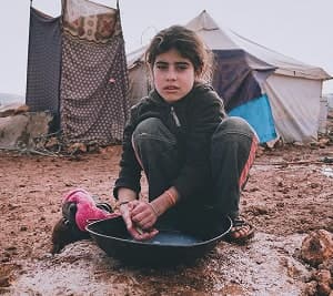 A young girl washing a bowl outside a tent in a muddy camp