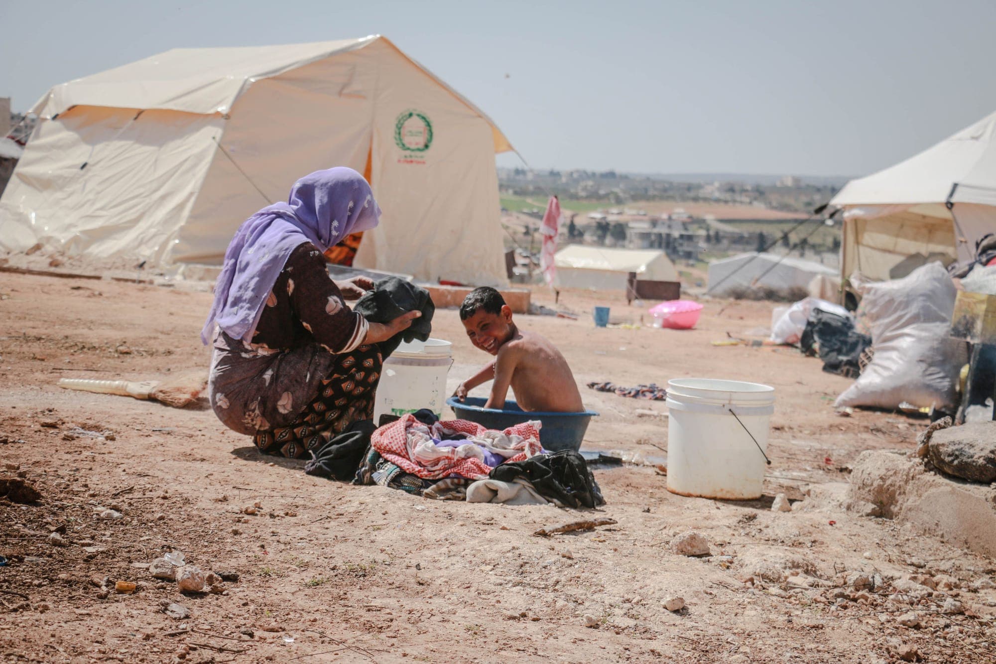 A mother washing clothes beside her child at a camp