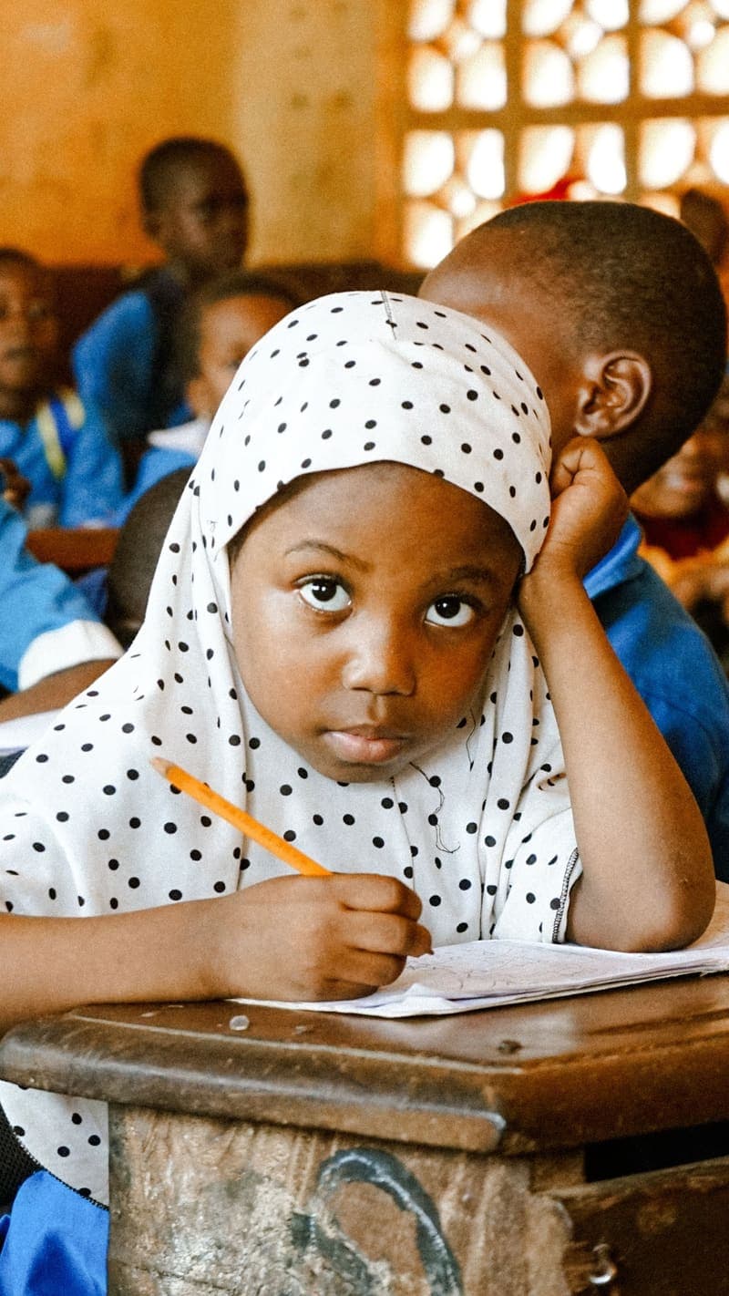 A young girl in a polka-dot hijab writing at a school desk